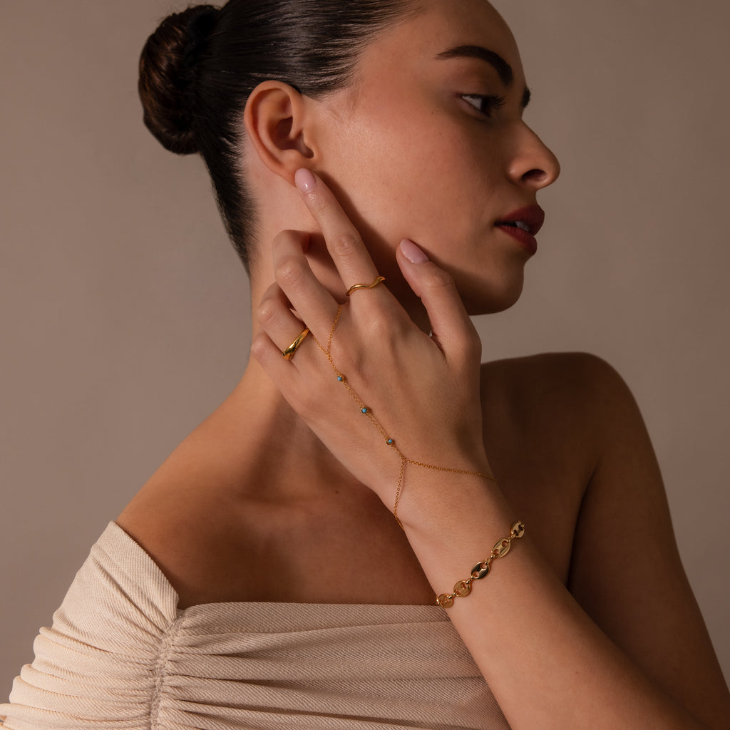 Woman in an off-shoulder top wears gold jewelry and the Turquoise Hand Chain Bracelet in 18K Gold, posing with her hand near her face against a neutral background.