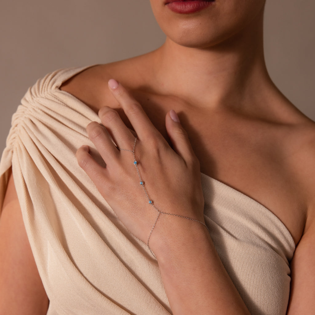 Woman in a cream off-shoulder dress wears the Turquoise Hand Chain Bracelet in Silver, adorned with turquoise stones, on her left hand.