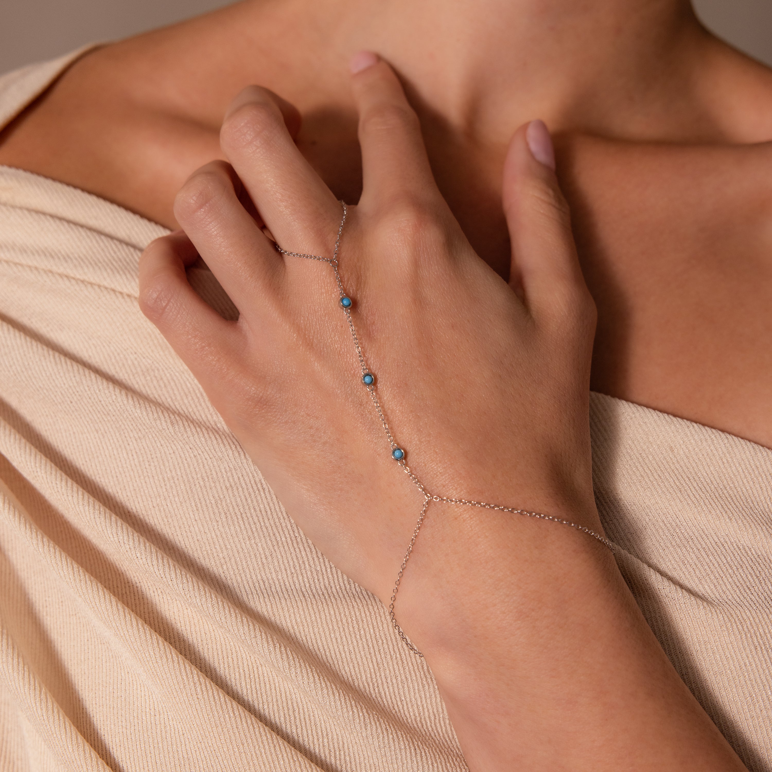 The Turquoise Hand Chain Bracelet in Silver, adorned with blue stones, elegantly graces a woman's hand as she rests it on a beige off-shoulder top.
