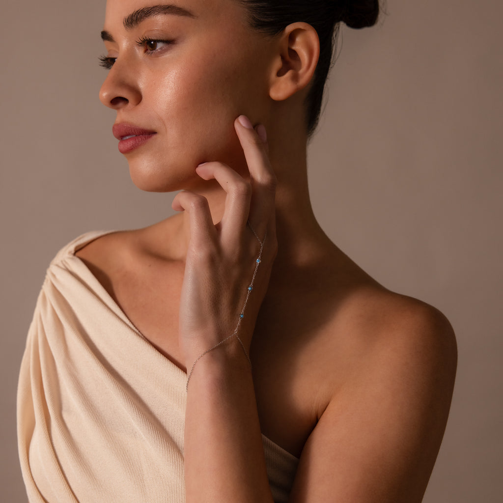 Woman in a cream off-shoulder dress touches her face, wearing the Turquoise Hand Chain Bracelet in Silver, and looks to the side.