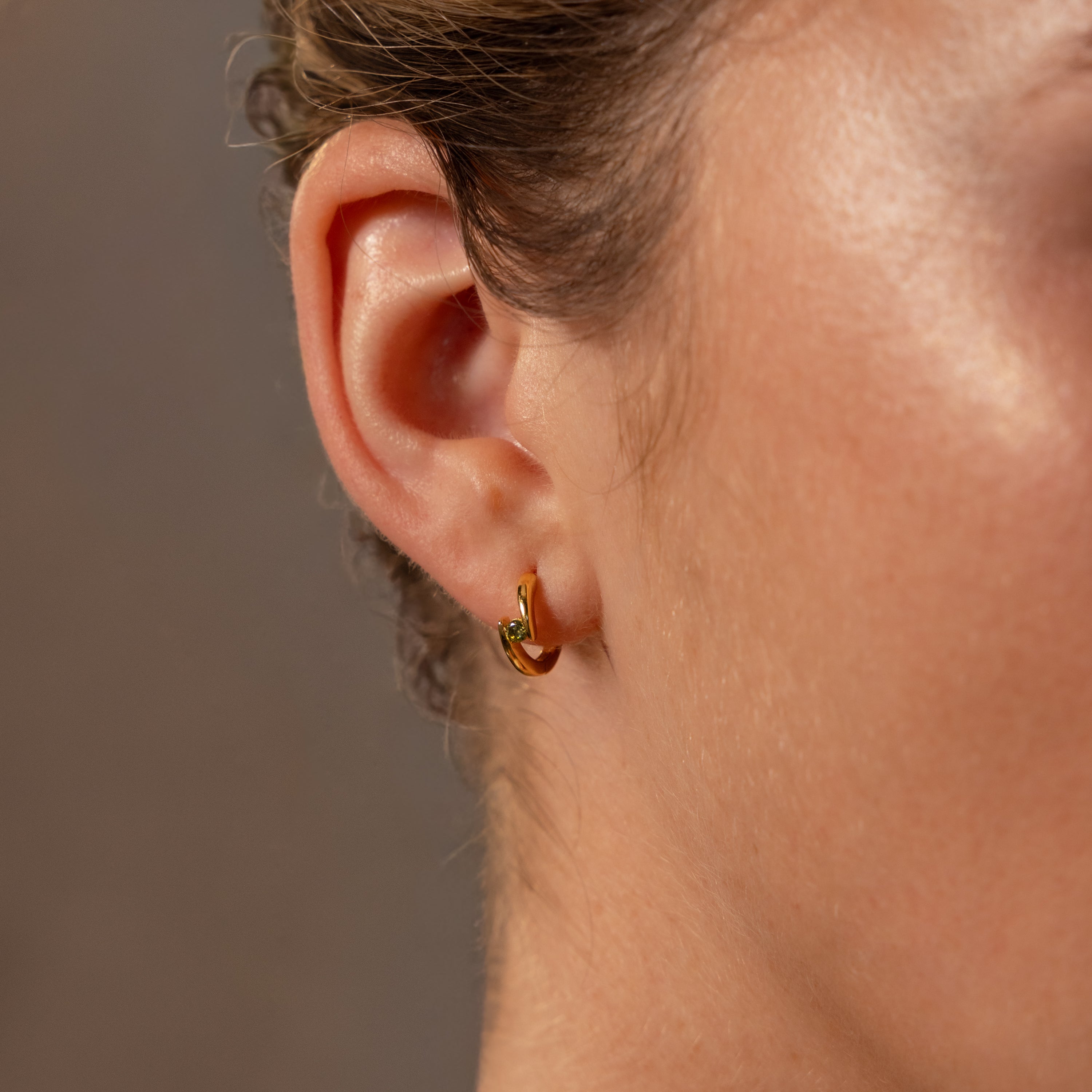Closeup of a womans ear wearing Floating Blue Zircon Huggies against a blurred brown background.