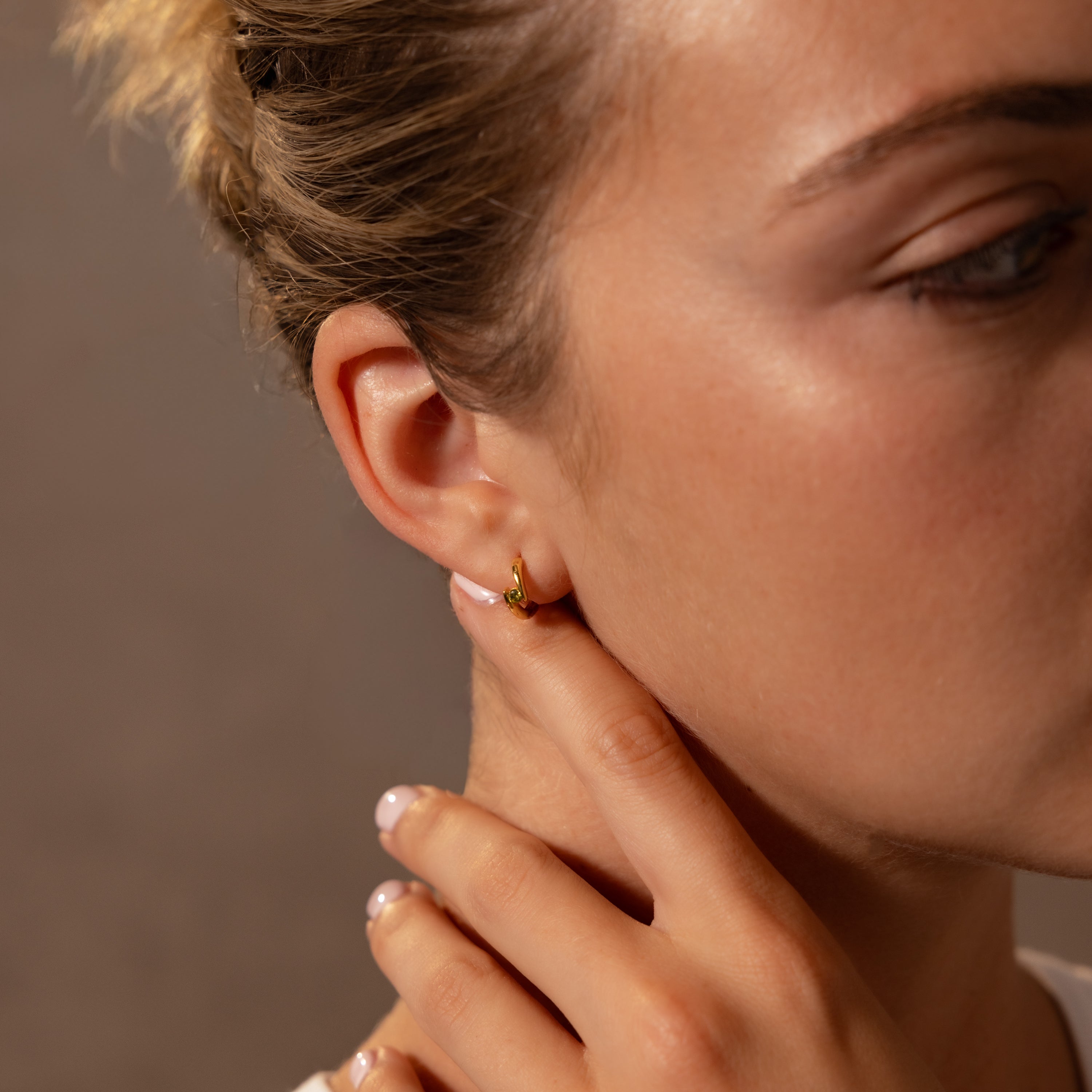 A close-up of a woman touching her neck, wearing Floating Birthstone Huggies—small gold hoop earrings—in her left ear.