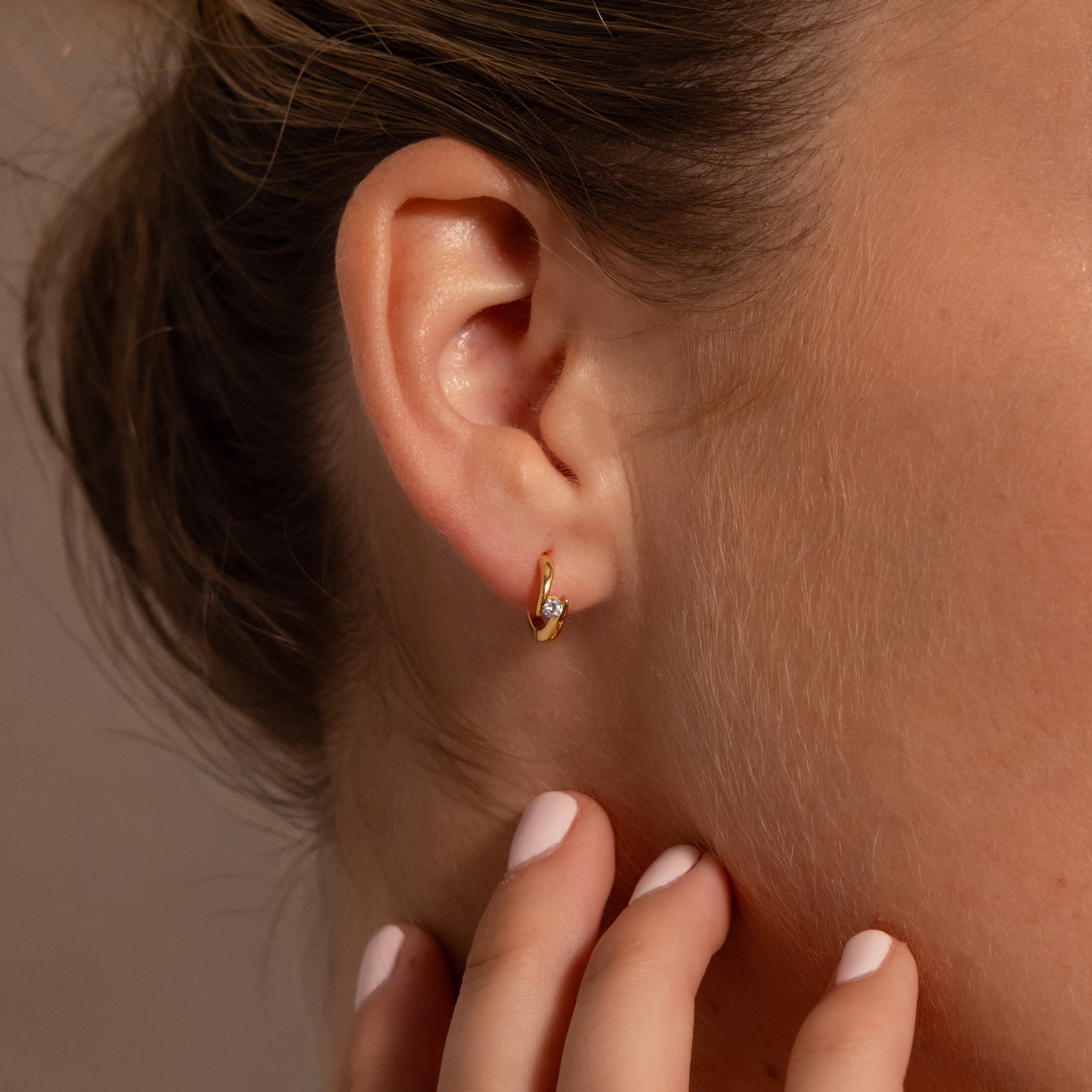 A close-up of a womans ear wearing Floating Diamond Huggies, with her hand, featuring pale pink nails, gently touching her neck.