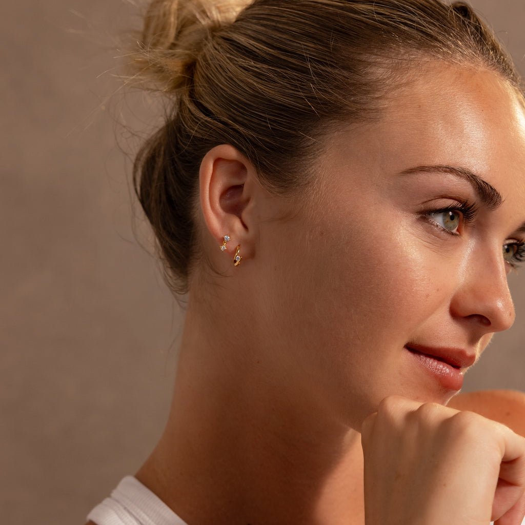 A woman with blonde hair in a bun, wearing Floating Topaz Huggies, gazes thoughtfully to the side, highlighting her personalized jewelry style.
