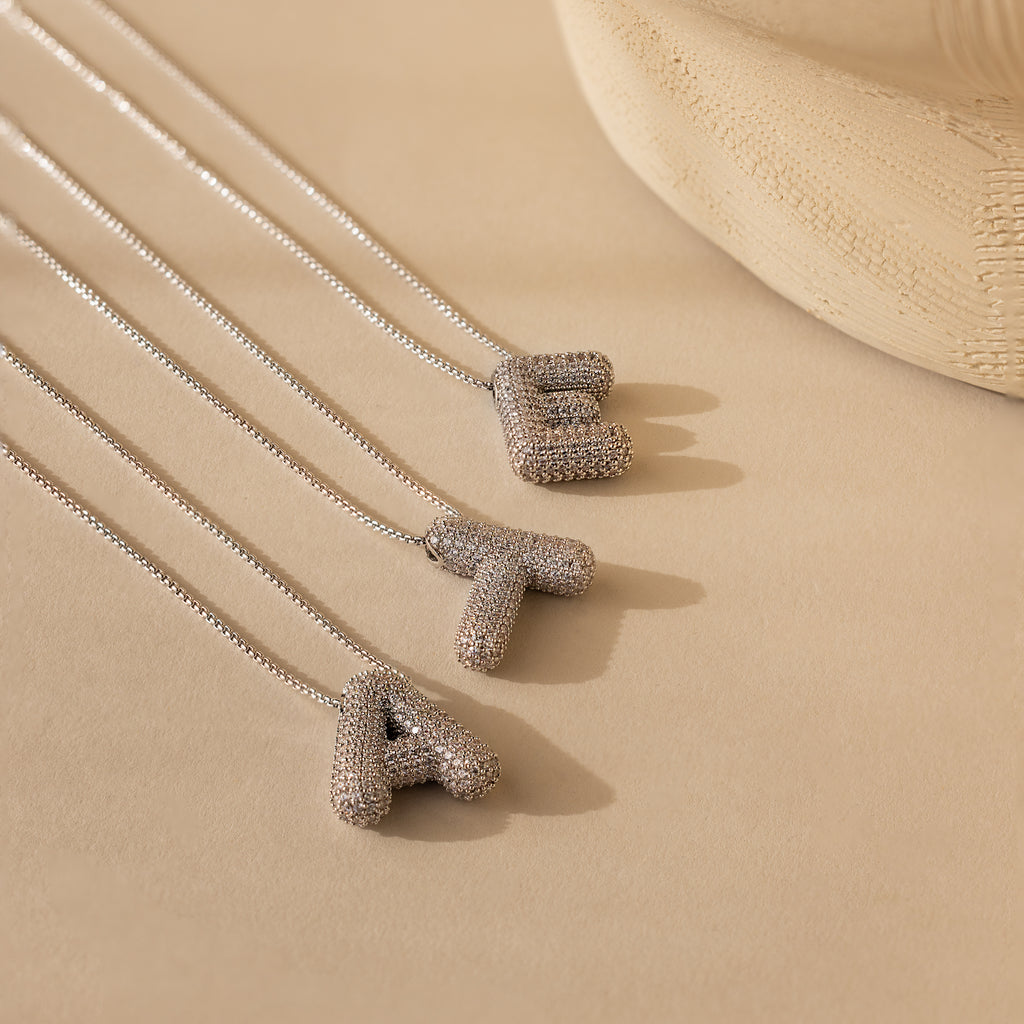 Three Pave Puff Letter Necklaces in White Gold—featuring textured pave A, T, and E pendants—rest on a beige surface.