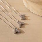 Three Pave Puff Letter Necklaces in White Gold—featuring textured pave A, T, and E pendants—rest on a beige surface.
