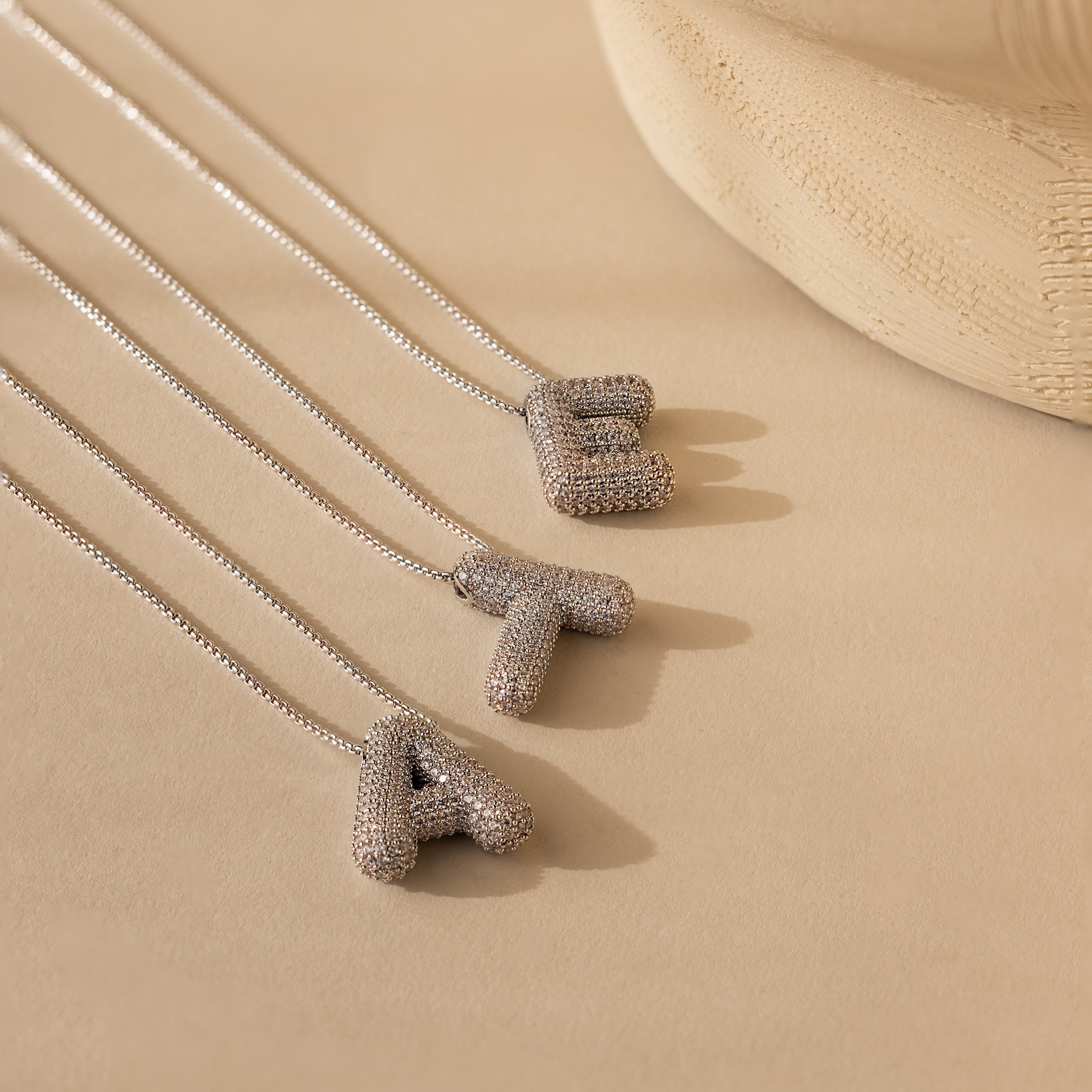 Three Pave Puff Letter Necklaces in White Gold—featuring textured pave A, T, and E pendants—rest on a beige surface.