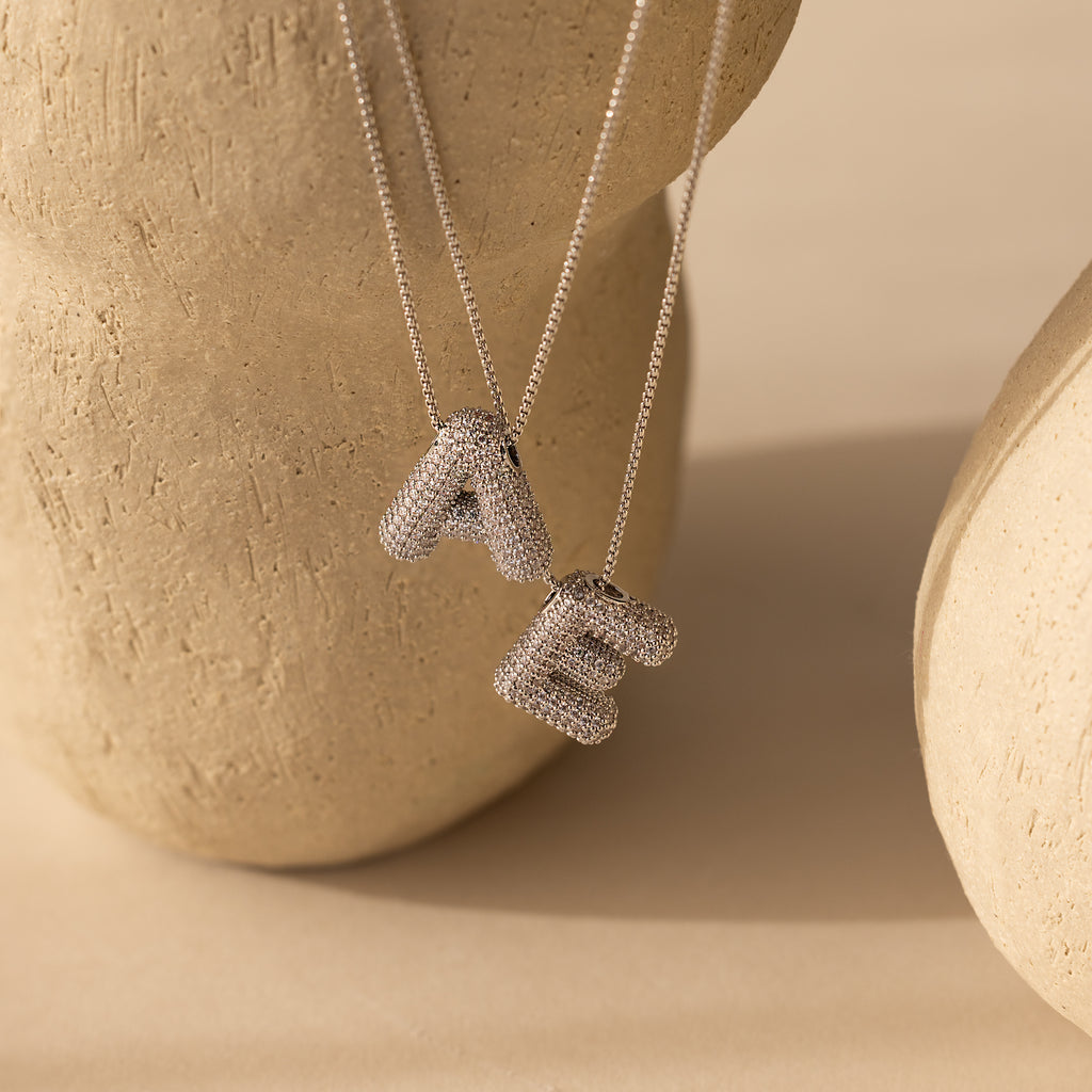 Two Pave Puff Letter Necklaces in White Gold, featuring textured A and E pendants with a chunky design, displayed on a beige textured background.