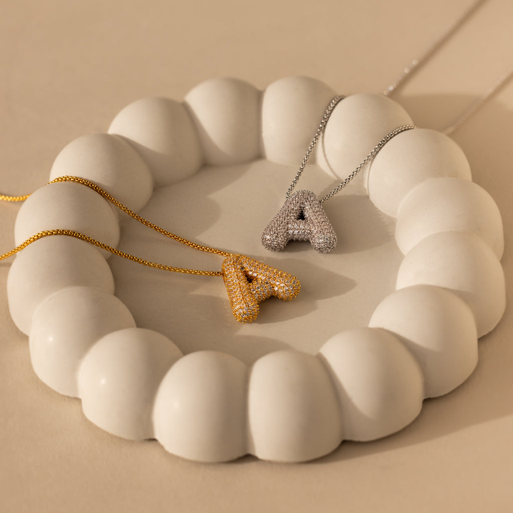 Two Pave Puff Letter Necklaces in 18K Gold, each with a sparkling initial pendant, are elegantly displayed on a white, round sculptural stand.