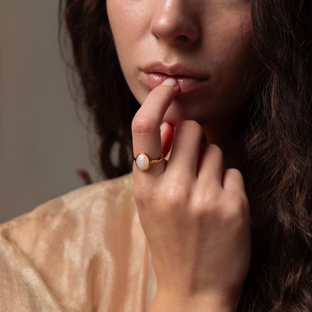 A woman with wavy brown hair thoughtfully touches her lips, wearing the Moonstone Signet Ring—an elegant everyday accessory.