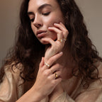 A woman with wavy brown hair poses with her hands near her face, thoughtfully looking down while wearing gold rings and the Moonstone Signet Ring.