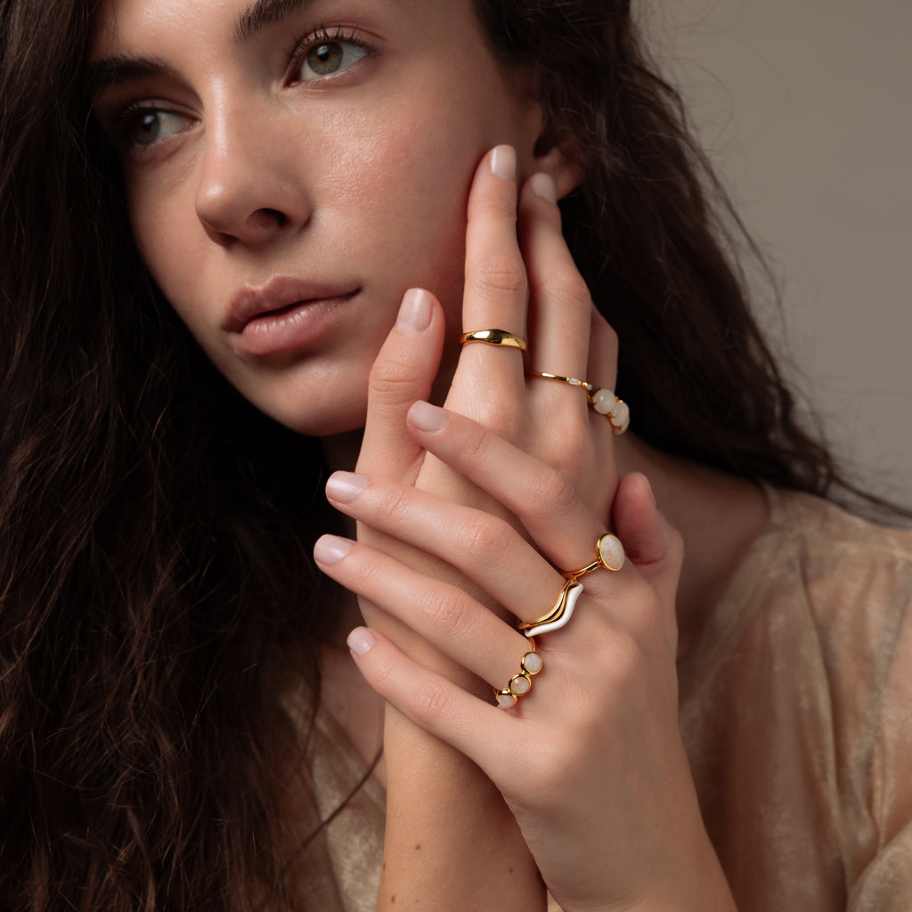 A woman with long brown hair thoughtfully touches her face and hands, wearing gold rings and the Moonstone Cluster Ring.