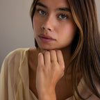 A woman with long brown hair wearing a Dainty Birthstone Drop Ring gazes softly, dressed in a light sheer top—a stunning piece of personalized jewelry.