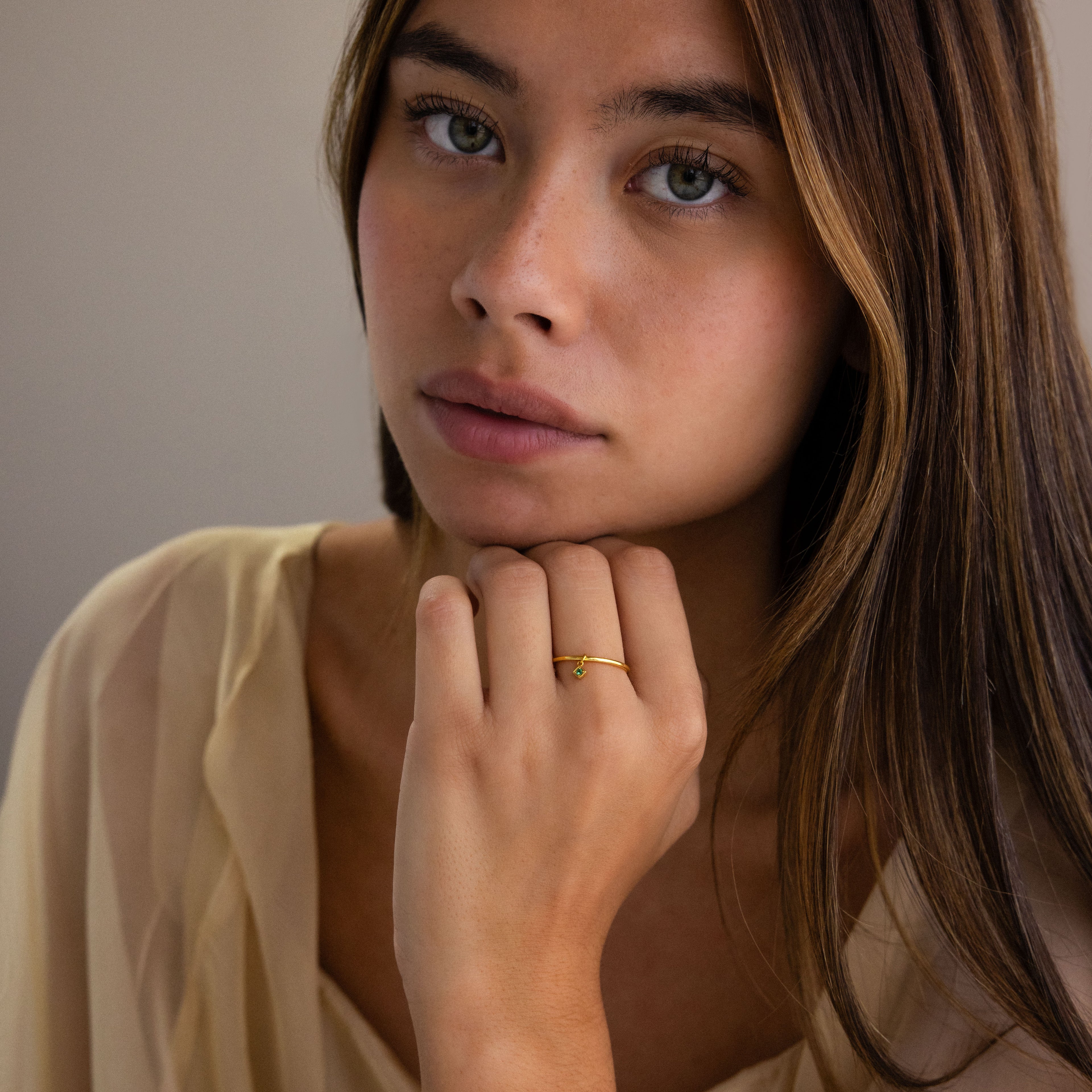 A woman with long brown hair wearing a Dainty Birthstone Drop Ring gazes softly, dressed in a light sheer top—a stunning piece of personalized jewelry.