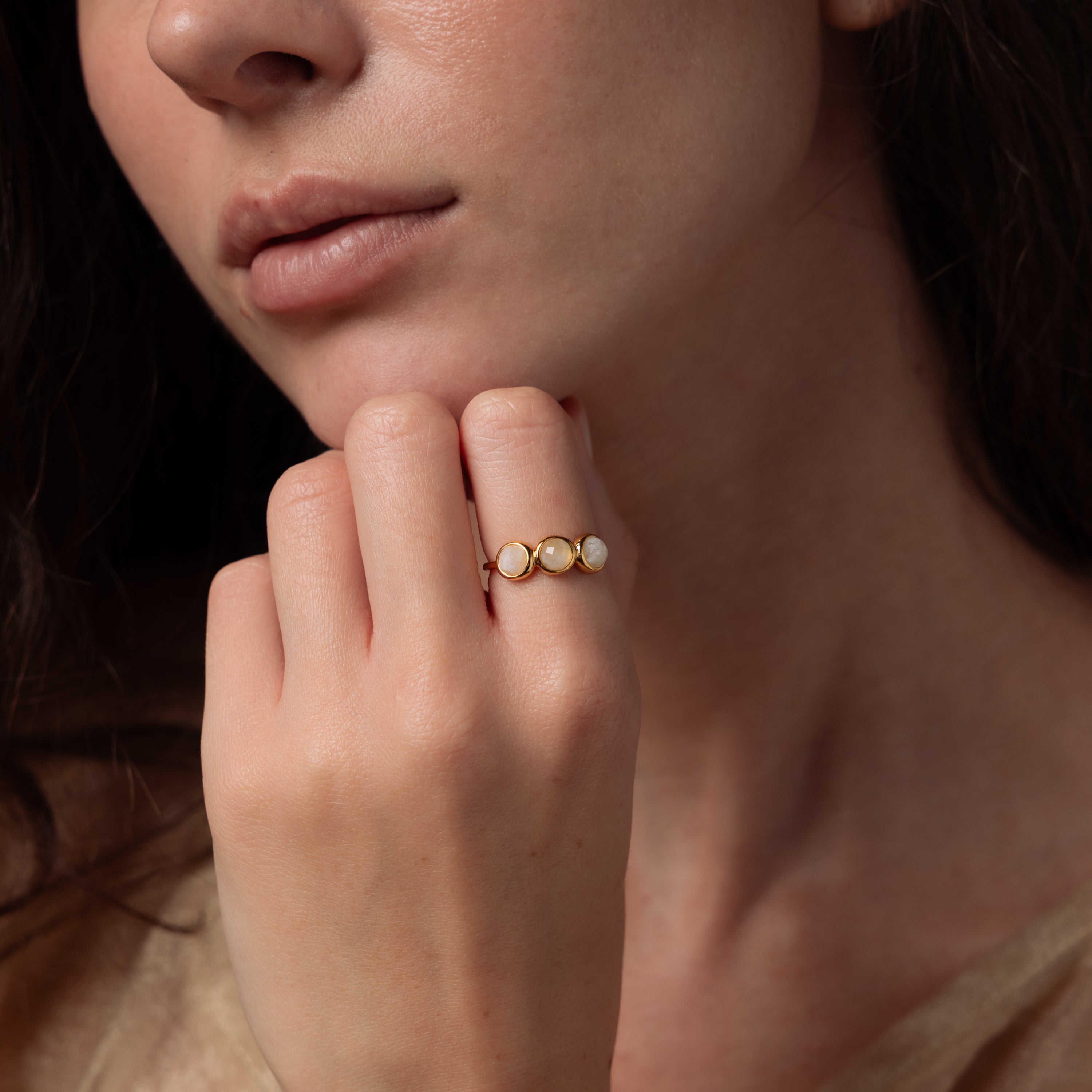 A woman's hand, near her face, displays the Moonstone Cluster Ring—a gold ring adorned with a moonstone and three round pearls.