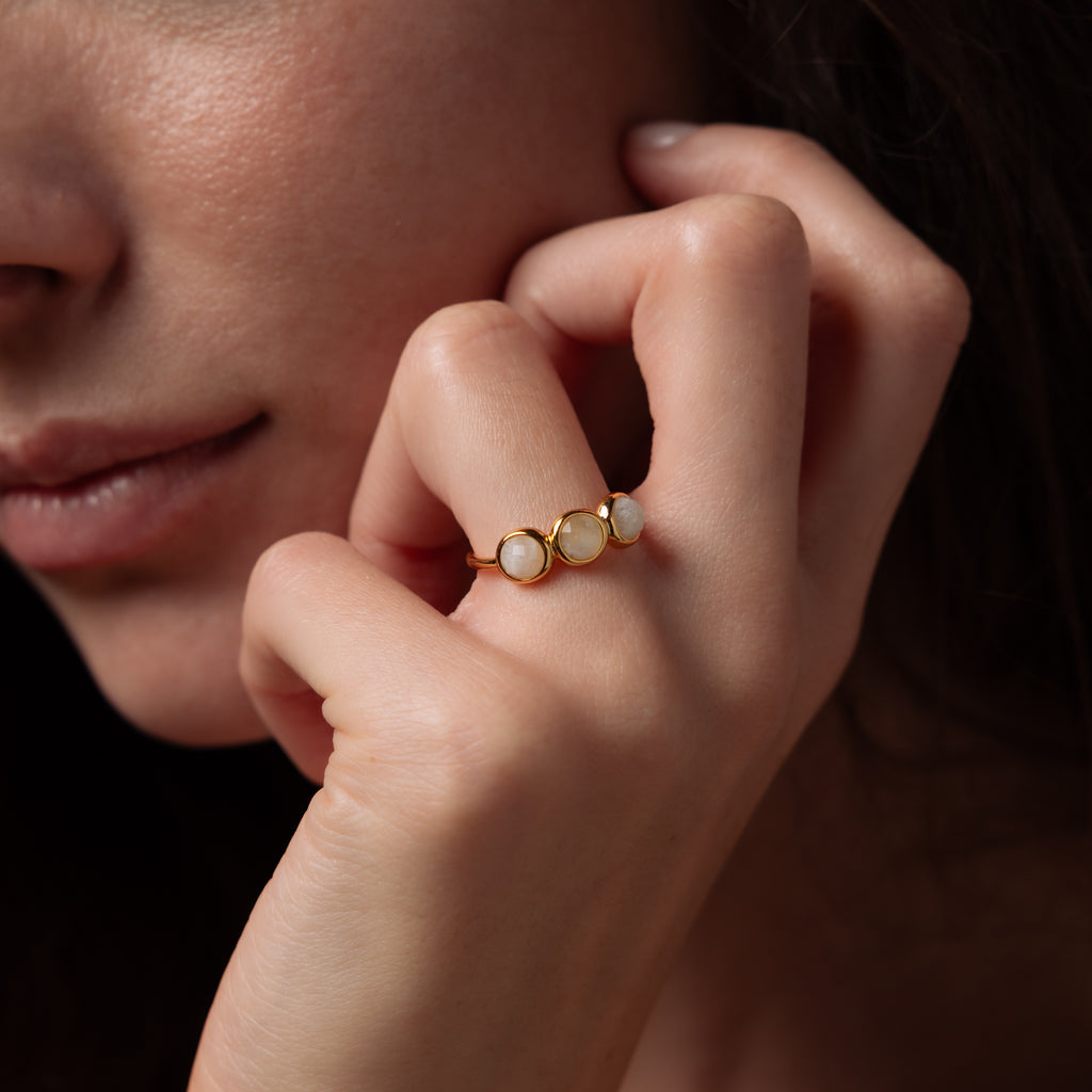 A close-up of a woman's hand near her face, adorned with the Moonstone Cluster Ring featuring three round white moonstones set in gold.