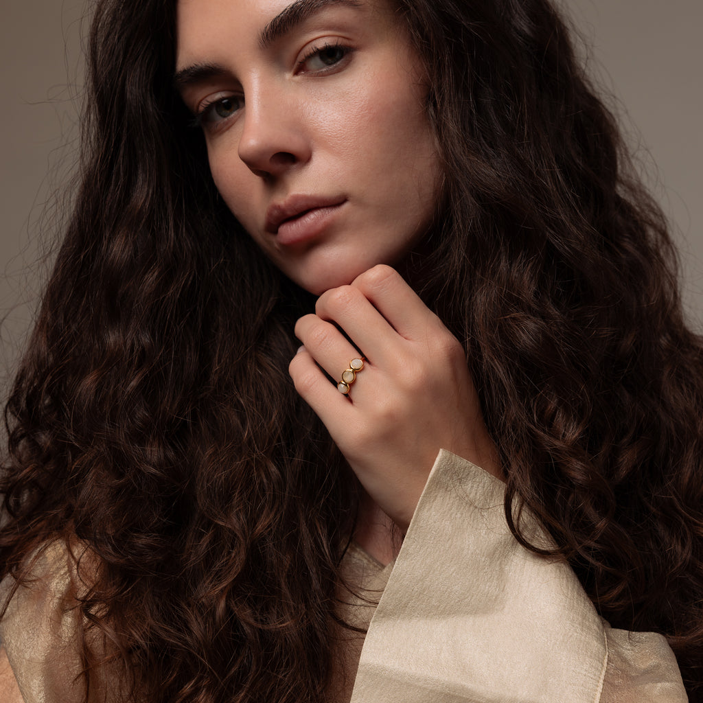 Woman with long curly hair wearing a beige top and gold rings, including the Moonstone Cluster Ring, poses with her hand near her face.