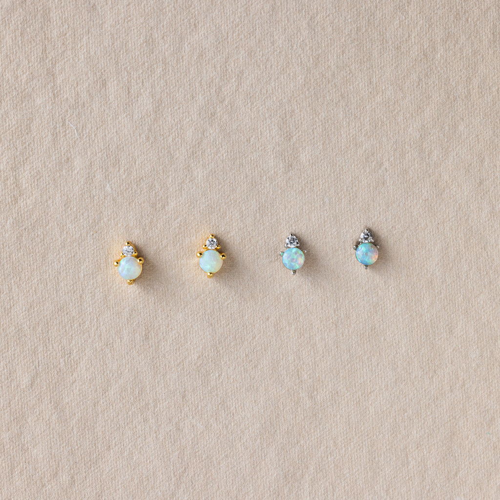 Four dainty Raindrop Emerald Studs with gold and silver settings are displayed on a textured beige surface.
