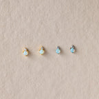 Four dainty Raindrop Emerald Studs with gold and silver settings are displayed on a textured beige surface.