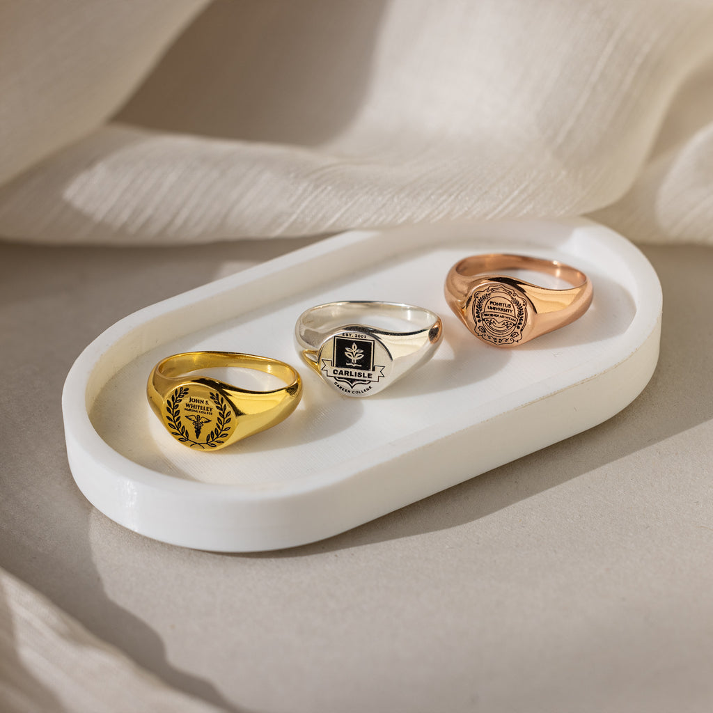 Three College Emblem Signet Rings in gold, silver, and rose gold are displayed on a white oval tray against a soft fabric background—an elegant graduation gift.