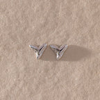 The Shark Tooth Flatback Studs in Silver rest on a textured beige surface—a stylish beach accessory to elevate your look.