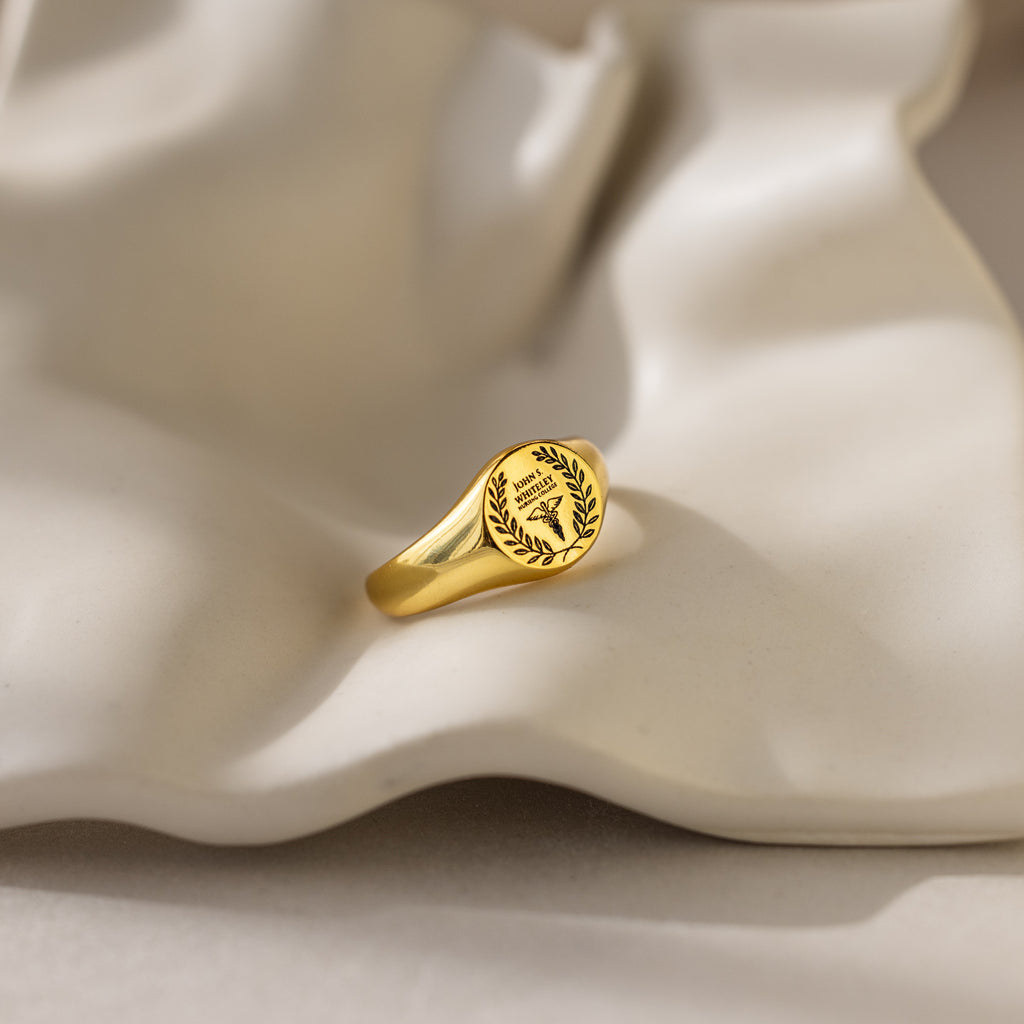 The College Emblem Signet Ring, ideal as a graduation gift or personalized accessory, is displayed on a wavy white ceramic surface.