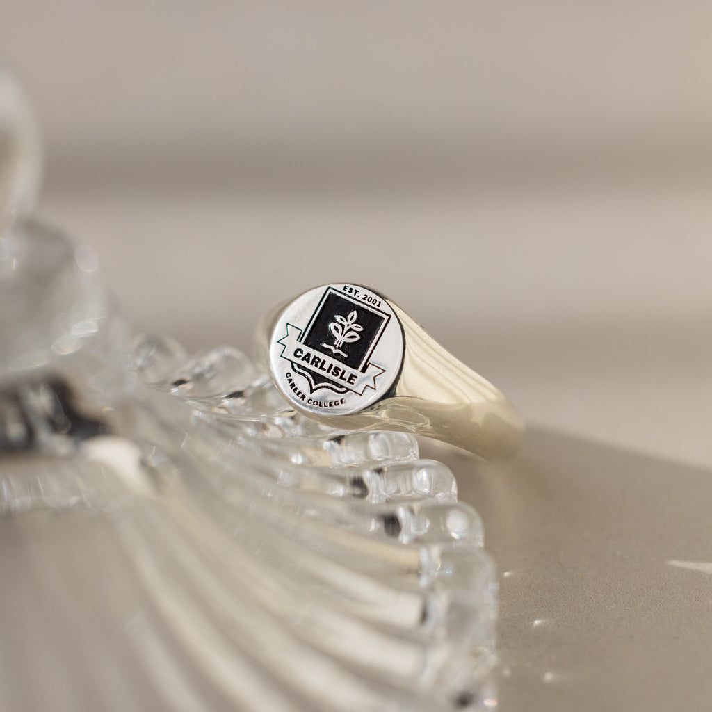 A silver College Emblem Signet Ring rests on a clear decorative glass surface, making it an ideal graduation gift.