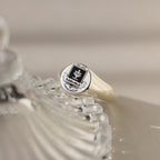 A silver College Emblem Signet Ring rests on a clear decorative glass surface, making it an ideal graduation gift.