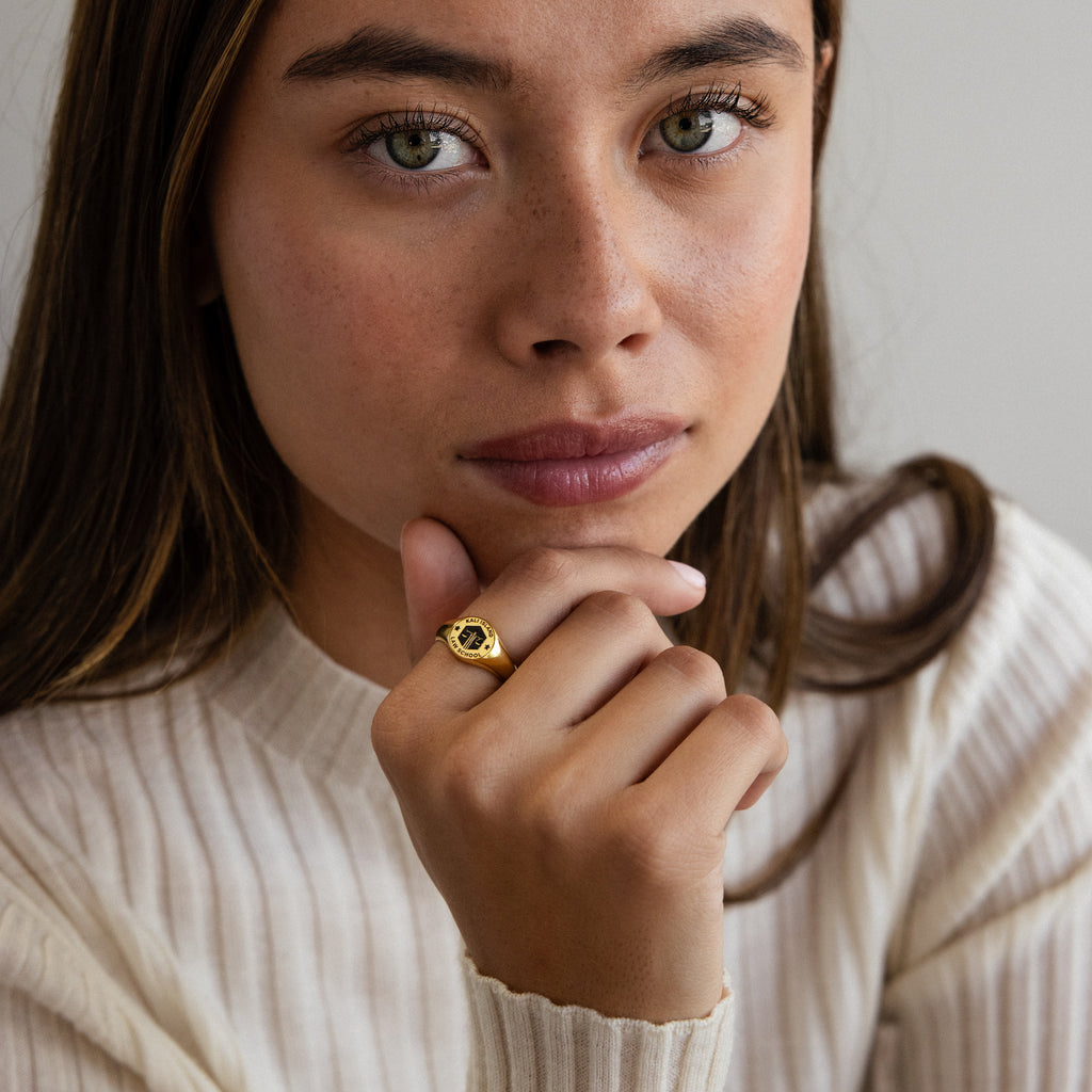 A woman with long brown hair in a cream sweater wears the College Emblem Signet Ring, gazing thoughtfully at the camera—an ideal choice for a meaningful graduation gift.