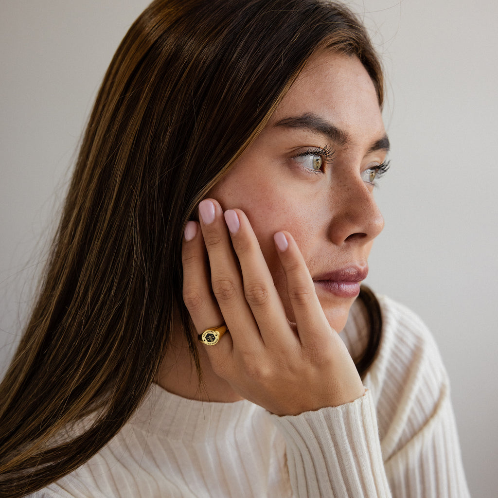 A woman with long brown hair rests her hand on her cheek, wearing the College Emblem Signet Ring—an elegant accessory and perfect graduation gift.