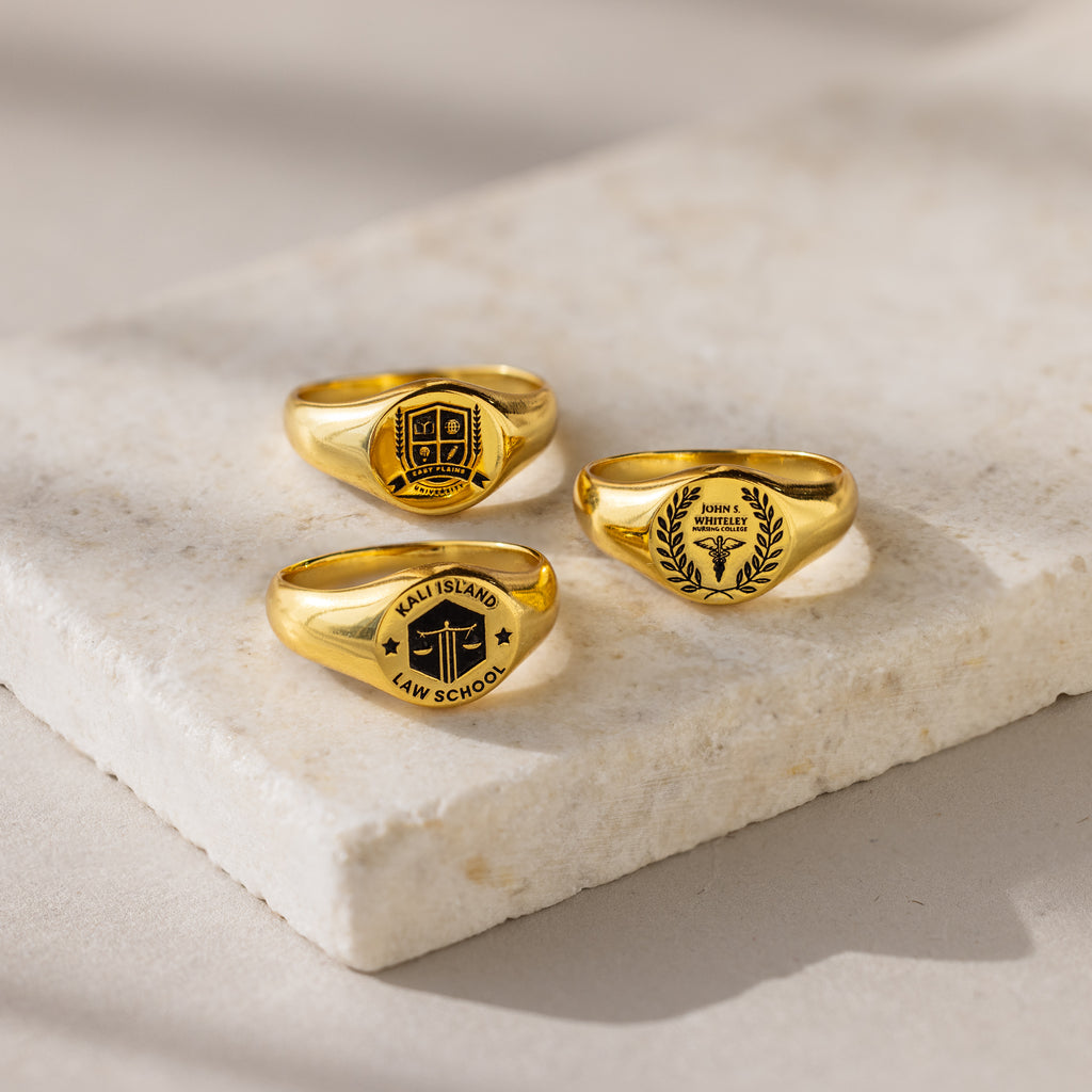 Three College Emblem Signet Rings with engraved law school emblems are displayed on a light stone surface—an ideal graduation gift to celebrate achievement.