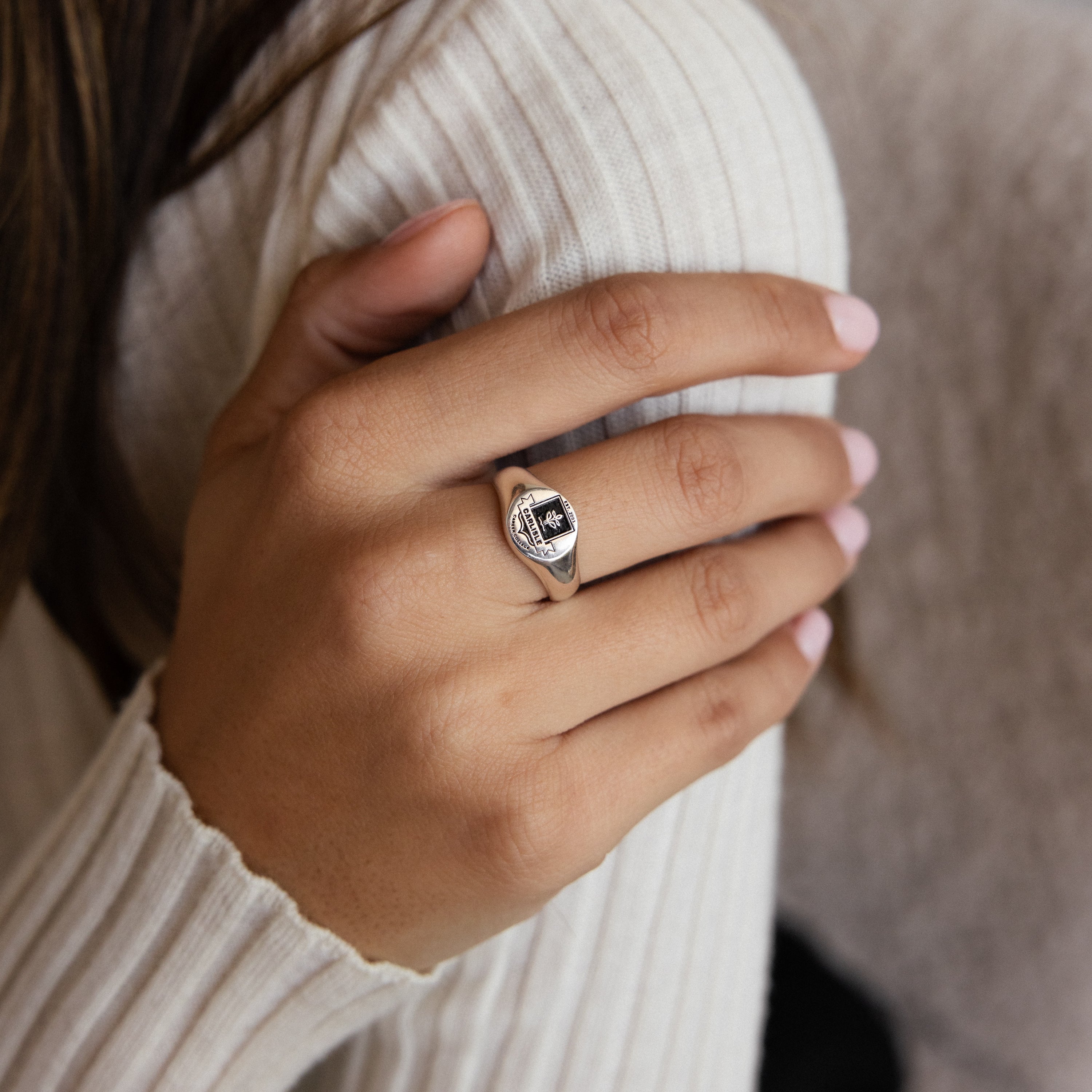 A hand with light pink nails wears the College Emblem Signet Ring, featuring a silver band, black stone, and clear accents—an ideal graduation gift.