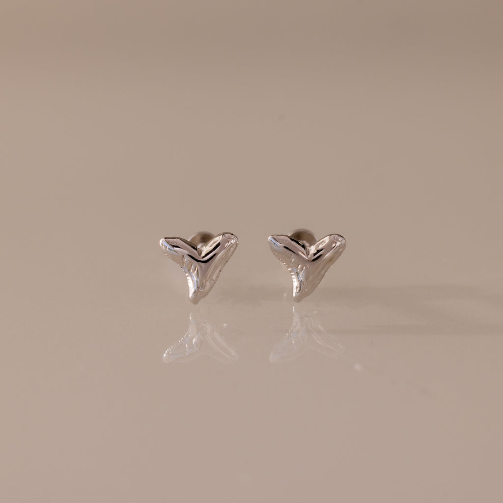 Pair of small, silver Shark Tooth Flatback Studs on a reflective light beige surface—an elegant beach accessory perfect for ocean lovers.