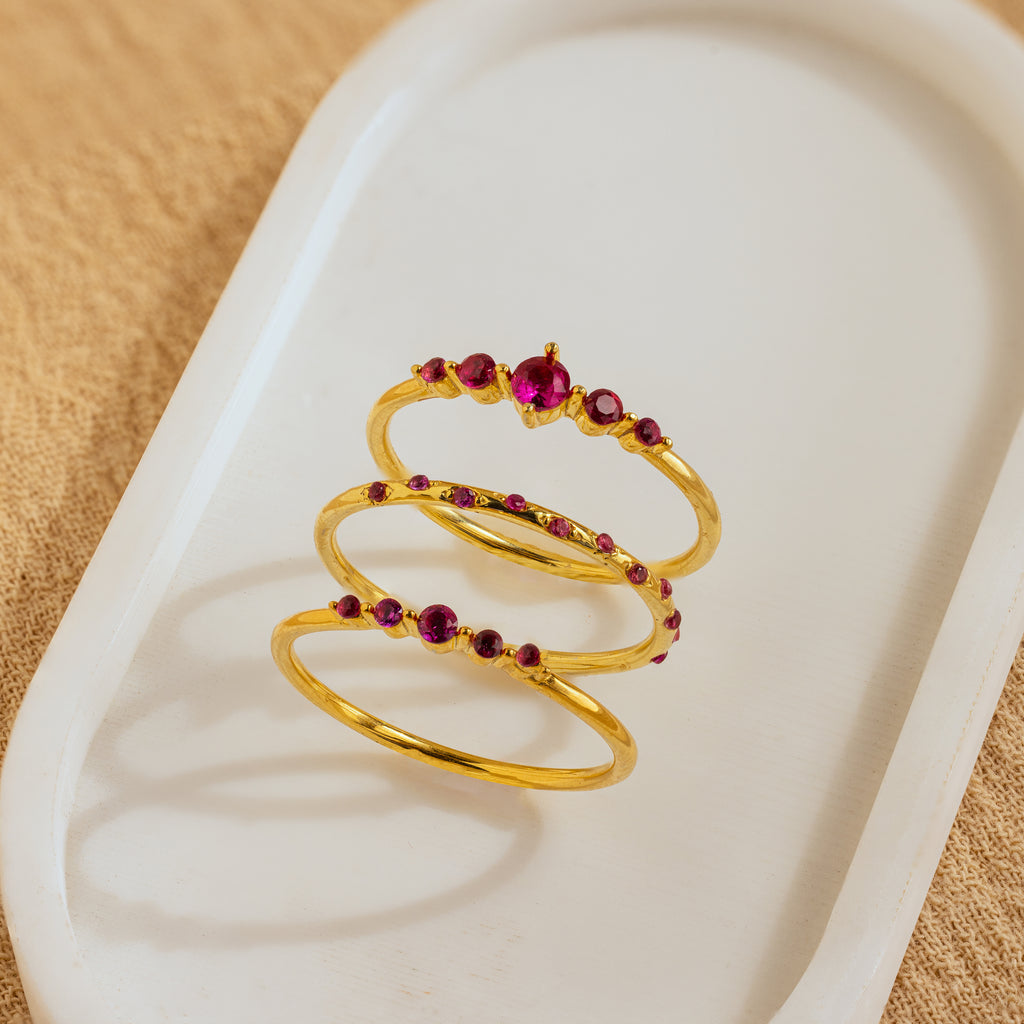 The Lace Birthstone Rings Set features three gold rings with red gemstones, elegantly displayed on a white oval tray with a beige fabric background—an ideal personalized gift.
