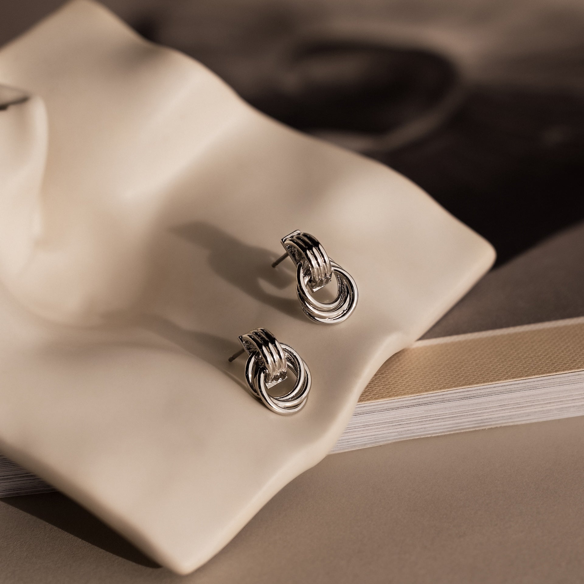 Micro Triple Knot Earrings in silver are displayed on a wavy ceramic dish with a book underneath, capturing the essence of quiet luxury.
