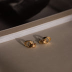 The Micro Triple Knot Earrings in 18K Gold rest on a beige surface, embodying quiet luxury with a book elegantly placed in the background.