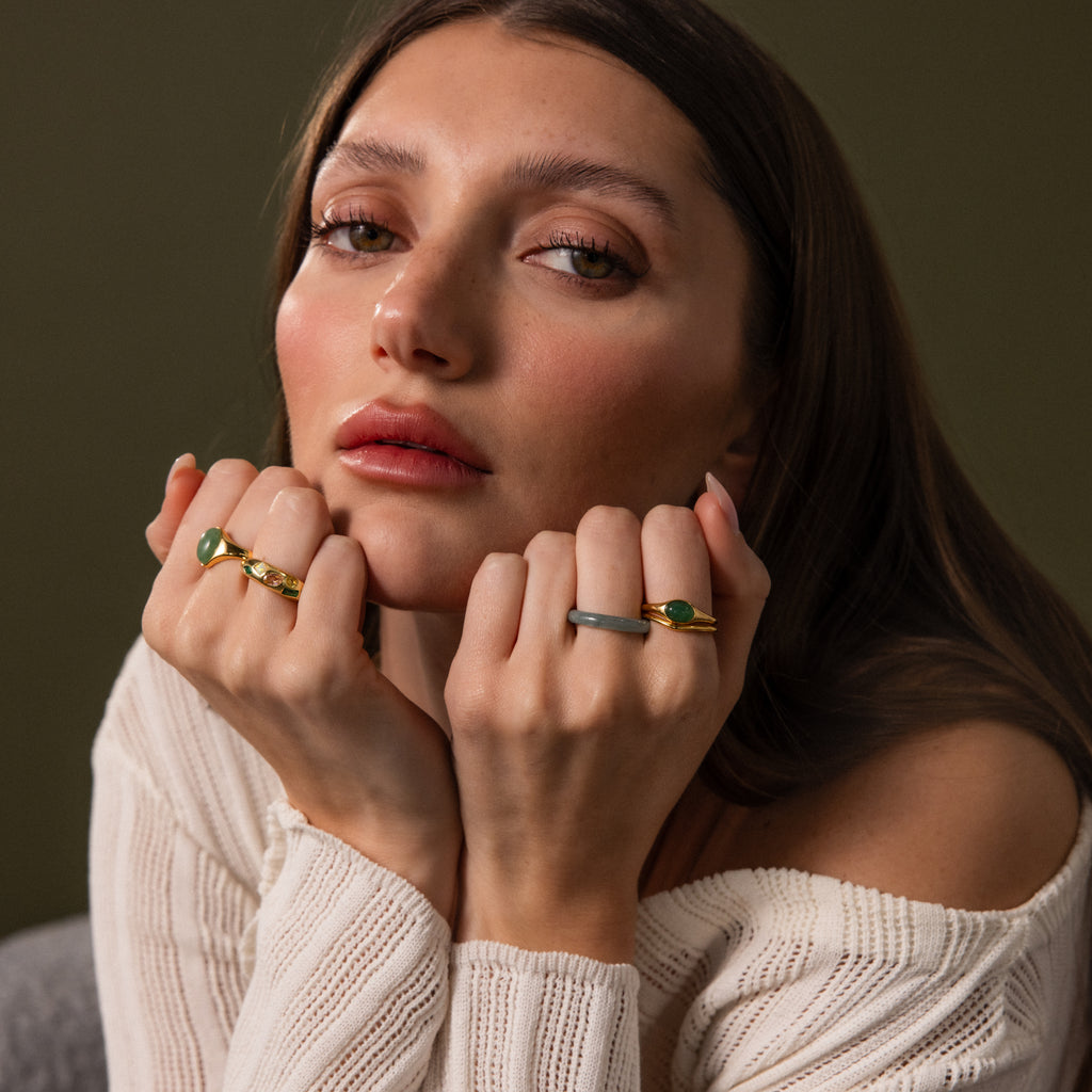 A woman with long brown hair, wearing elegant jewelry and an off-shoulder knit top, poses with hands near her face to showcase the Oval Jade Signet Ring.