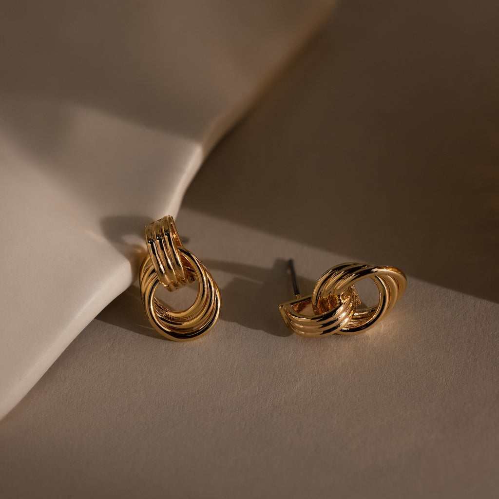 Micro Triple Knot Earrings displayed on a beige surface, with one earring lying flat and the other propped against a white object—an elegant touch of quiet luxury.