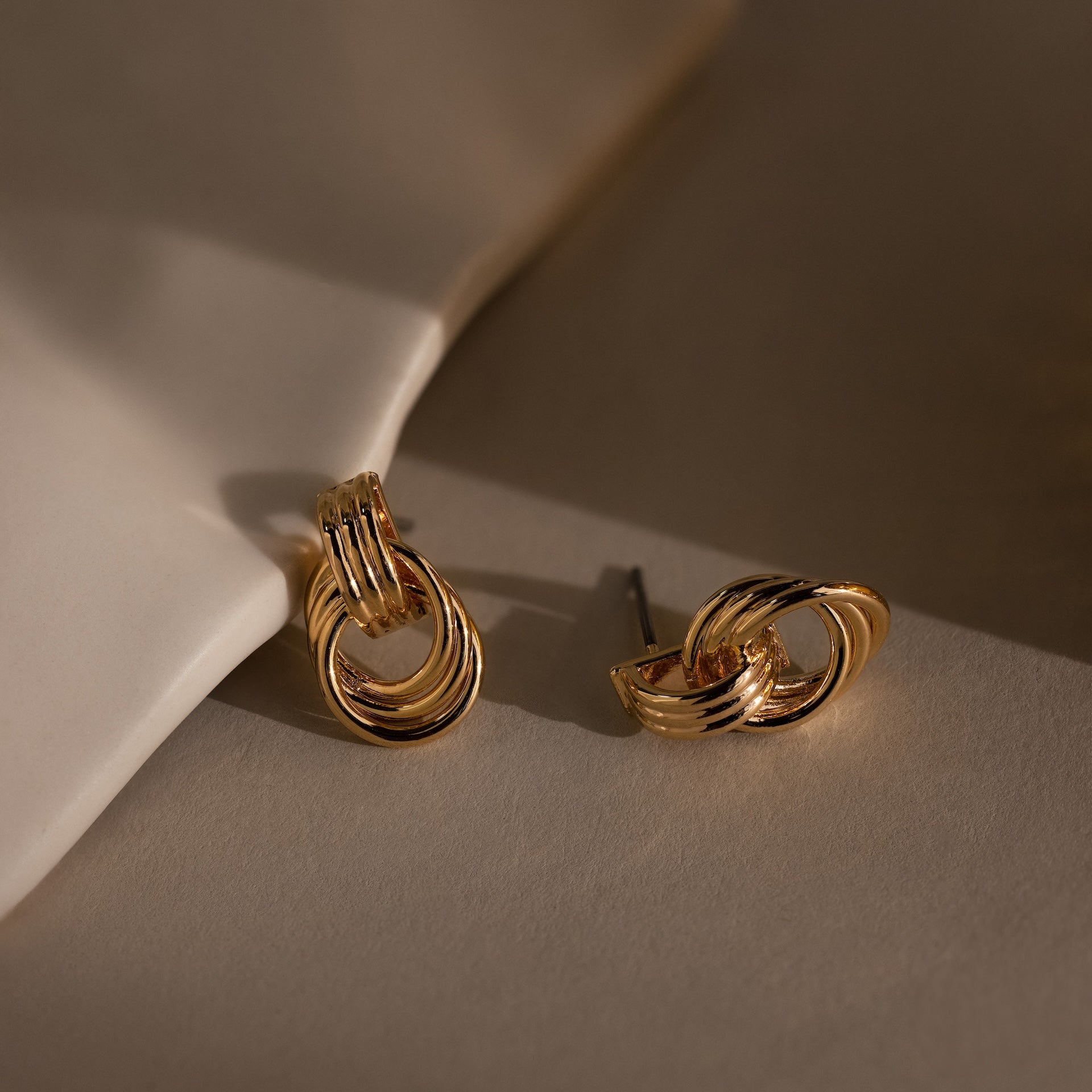 Micro Triple Knot Earrings displayed on a beige surface, with one earring lying flat and the other propped against a white object—an elegant touch of quiet luxury.
