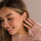 A smiling woman with wavy hair showcases Jade Marquise Huggies, pairing them with a sheer ruffled top and a gold ring.