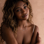 A woman with wavy hair wearing a white top showcases the Heart Station Necklace, featuring heart charms on a cable chain, against a beige background.