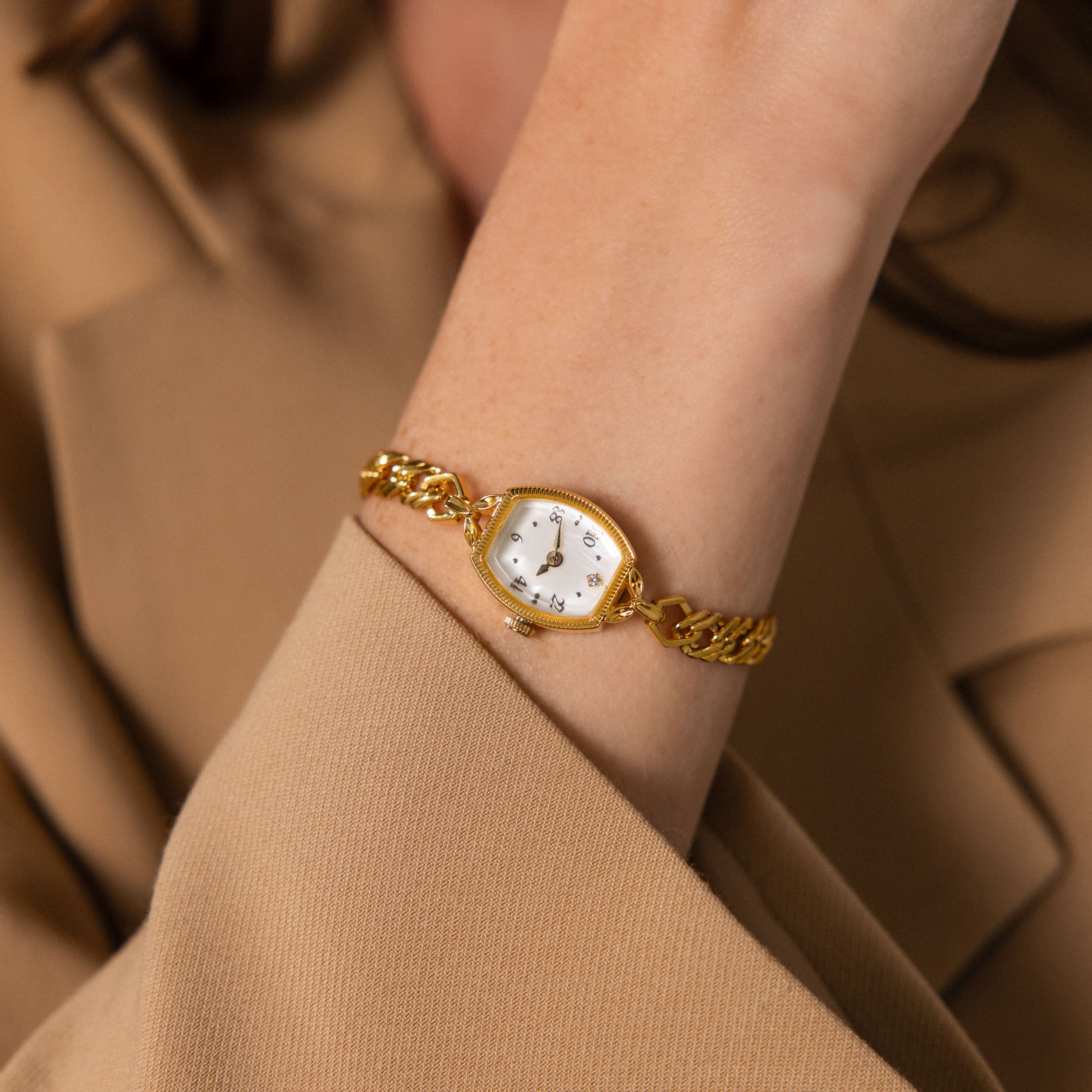 Heirloom Chain Link Timepiece featuring a gold chain band, white face, and diamond accents, shown on a person in a tan blazer with arm slightly raised.