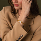 A woman in a beige blazer touches her face, wearing the Heirloom Chain Link Timepiece—a gold watch with a white dial.