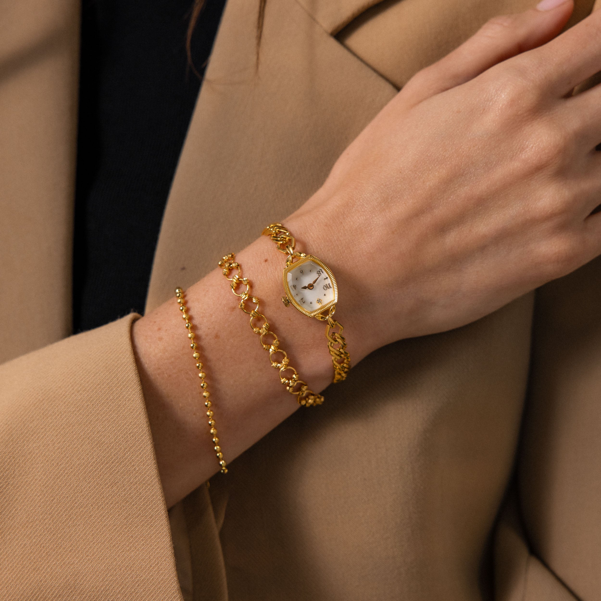 A person wearing the Heirloom Chain Link Timepiece and two gold bracelets, dressed in a tan blazer and black top.