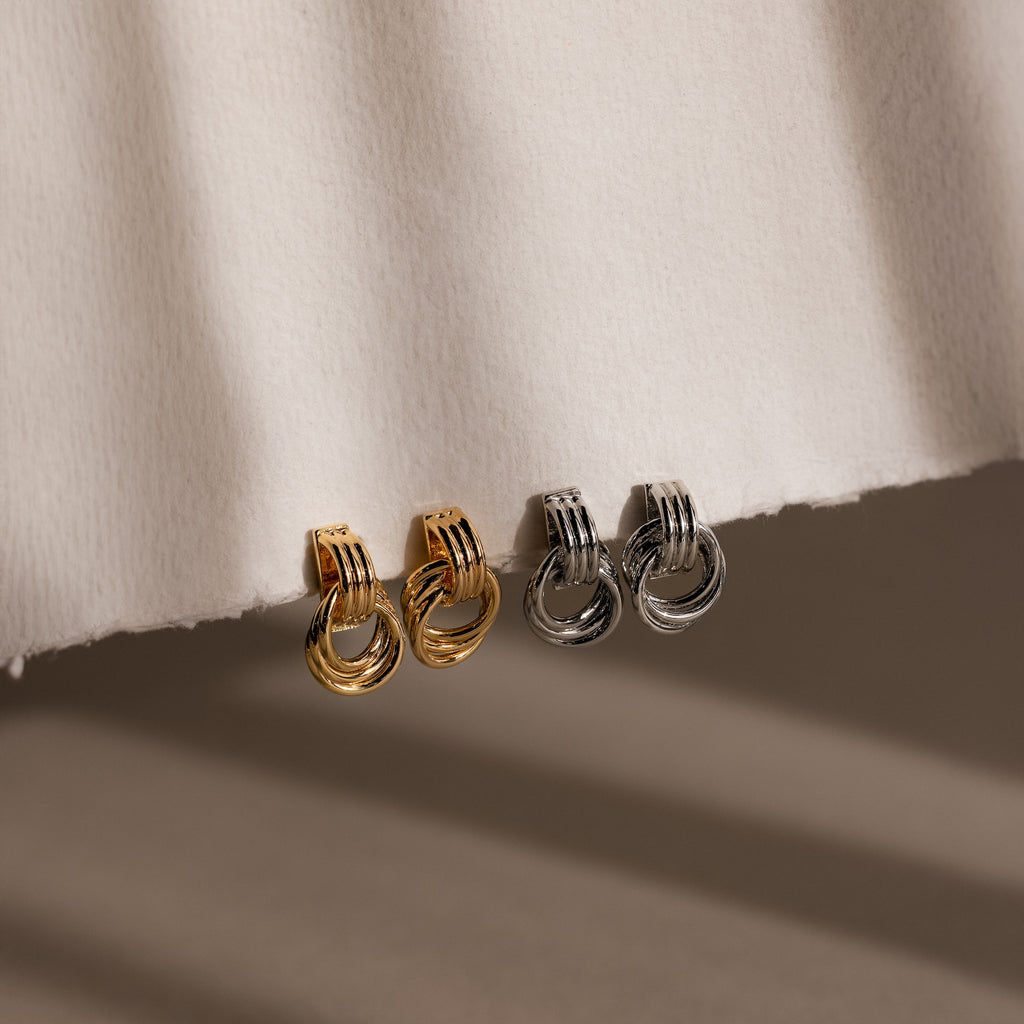 Micro Triple Knot Earrings in gold and silver, showcased on a textured white surface with gentle shadows, radiate quiet luxury.