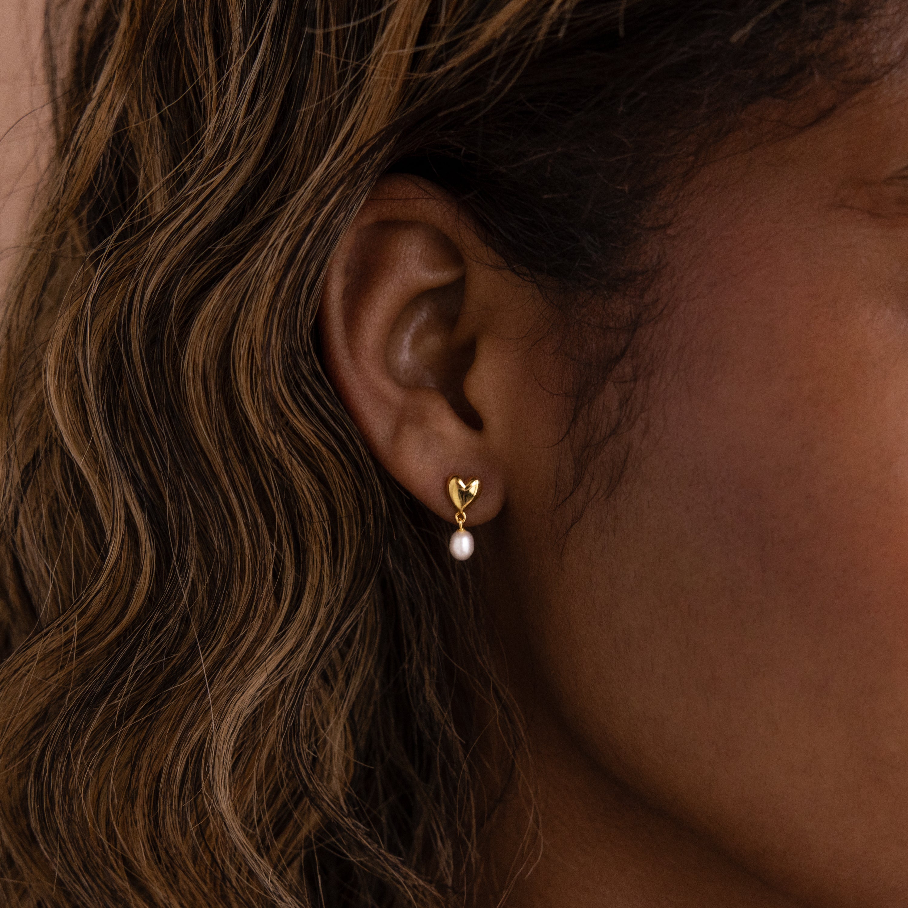 A womans ear wearing Heart Pearl Drop Earrings—gold heart studs with a dangling pearl.