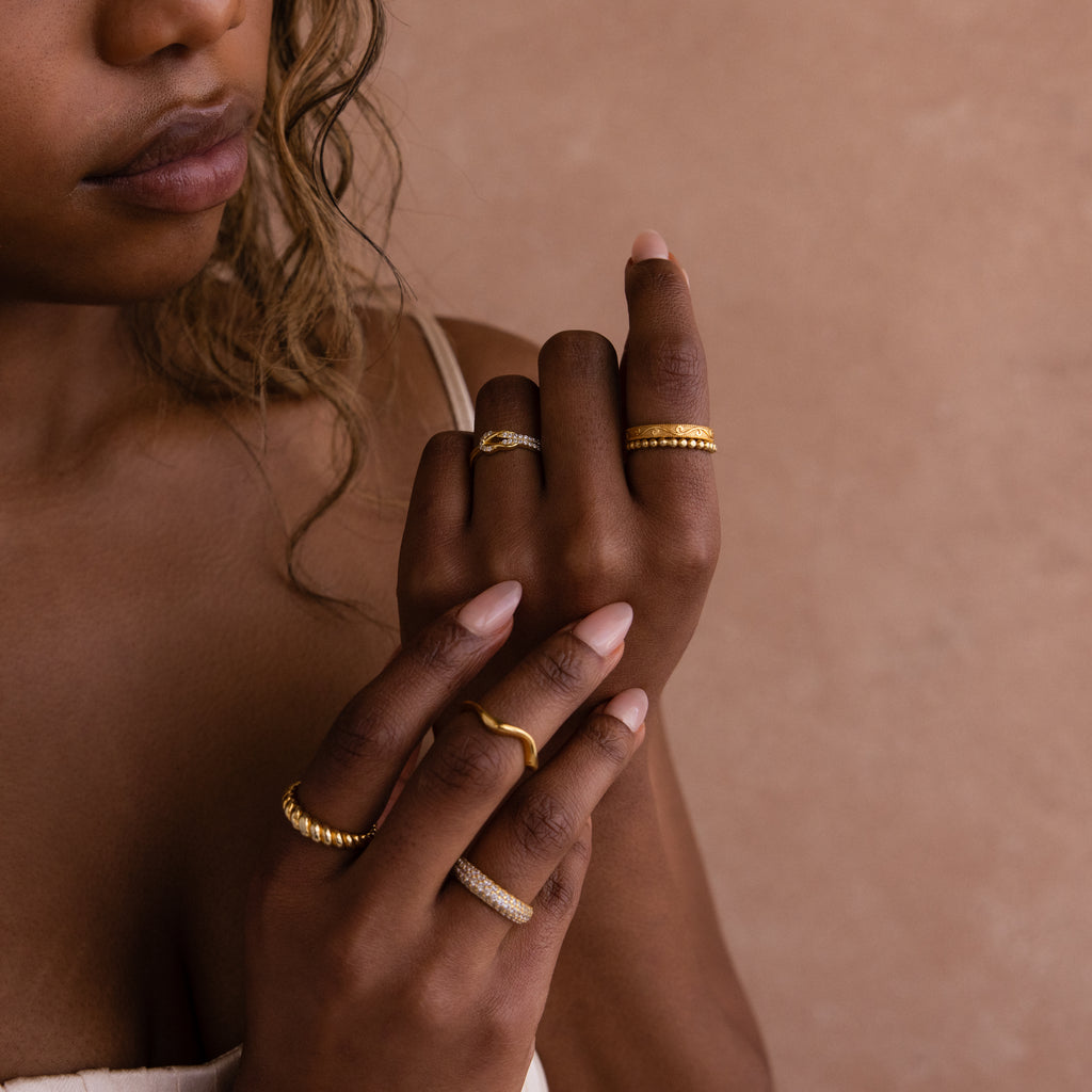 A woman with wavy hair displays her hands adorned with multiple gold rings, featuring the Pave Double Knot Ring, against a beige background.