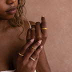 A woman with wavy hair displays her hands adorned with multiple gold rings, featuring the Pave Double Knot Ring, against a beige background.
