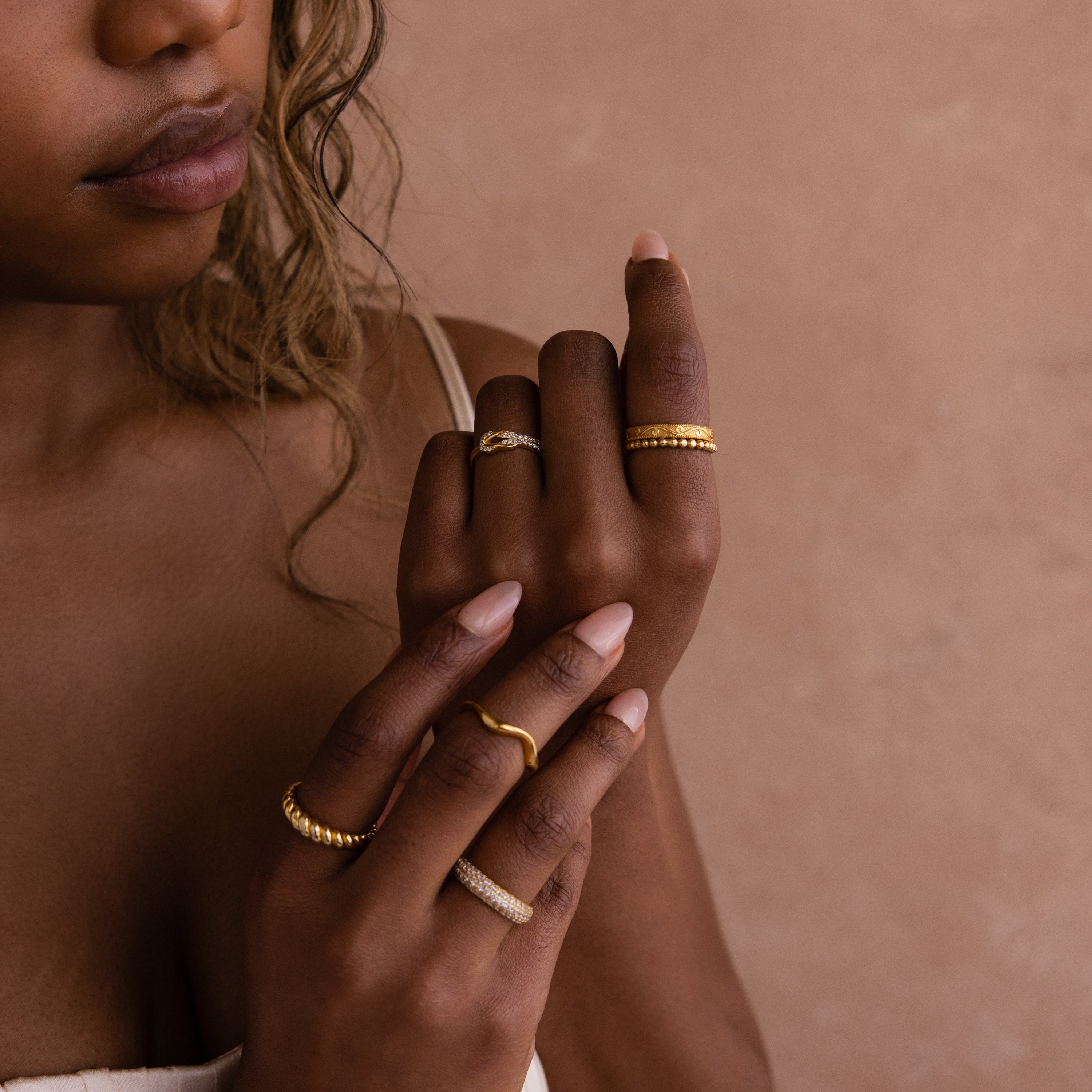 A woman with wavy hair displays her hands adorned with multiple gold rings, featuring the Pave Double Knot Ring, against a beige background.