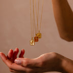 The Topaz Birth Flower Charm Necklace is displayed above an outstretched hand against a neutral, minimalist background—perfect for showcasing elegant jewelry.